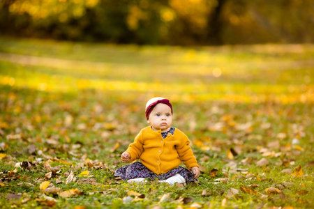 Baby girl in yelloy jacket and red headband seating on the grass, playing in the autumn leaves.