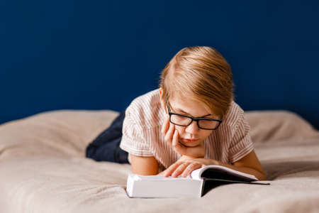 A 10-year-old boy with glasses is lying on the bed reading a big book.の写真素材
