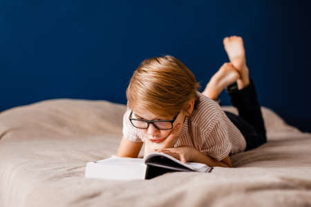 A 10-year-old boy with glasses is lying on the bed reading a big book.の写真素材
