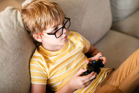 Blonde boy in big glasses playing on the console sitting on the couch. European child having a hobby.の写真素材