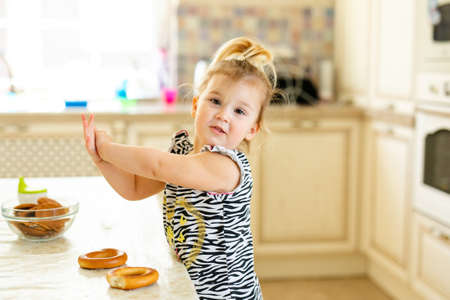 Little toddler kid having lunch in the warm sunny kitchen. Blonde girl with funny ponytail playing with two tasty bagels.の写真素材