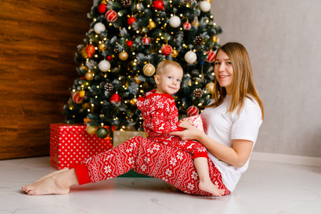 young mother and her toddler in red pajamas are joyfully playing by the Christmas tree.