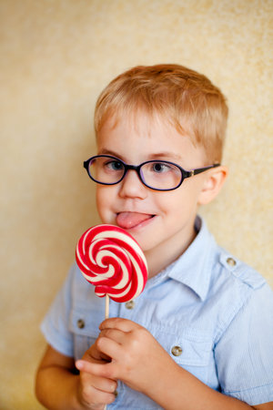 A young boy with glasses and squinting eyes is delightfully munching on a big red lollipopの写真素材