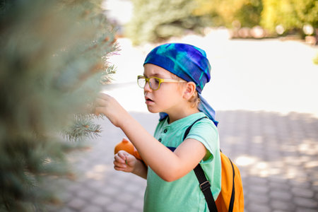 small boy in glasses and a headband, dressed in a green shirt, observing a plant with curiosity, holding a toy monkey in his handの写真素材