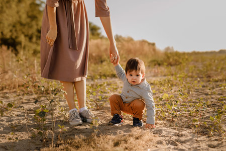 Mom holds her one-year-old baby by the hand for a walk in the park by the river.