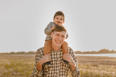 A child on his fathers neck. Walk near the water. Baby and dad against the sky.