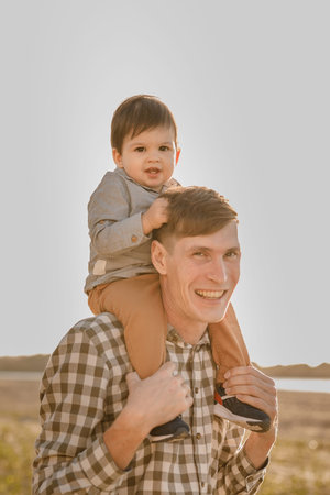 A child on his fathers neck. Walk near the water. Baby and dad against the sky.