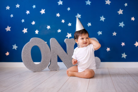 Baby boy of one year old wearing a white body and a festive paper hat sits on the floor on a blue background with stars and big silver letters.の写真素材