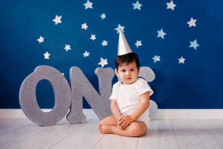 Baby boy of one year old wearing a white body and a festive paper hat sits on the floorの写真素材