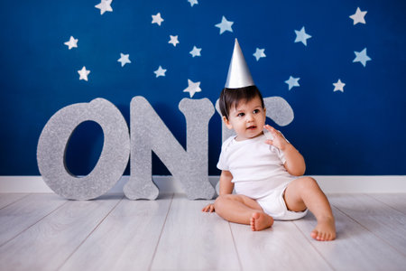 Baby boy of one year old wearing a white body and a festive paper hat sits on the floor.の写真素材