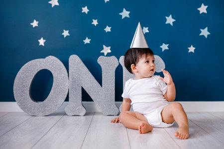 Baby boy of one year old wearing a white body and a festive paper hat sits on the floor on a blue background with starsの写真素材