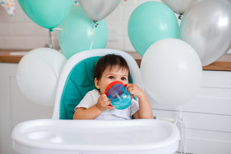 Little baby boy sitting in blue high chair at home on white kitchen and playing with wooden big spoon on background with balloonsの写真素材