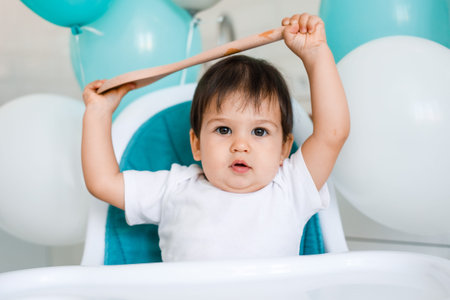 A one-year-old boy is seen in a high chair with a wooden spoon in his hands.