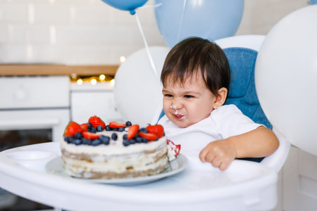 Little baby boy sitting in high chair in white kitchen and tasting first year cake with fruits on background with balloons
