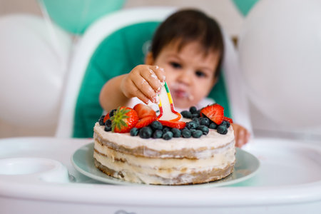 Little baby boy sitting in high chair in white kitchen and tasting first year cake with fruits on background with balloonsの写真素材