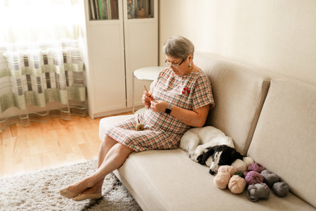 Old woman in glasses crocheting at home with her white dog next to her.の写真素材