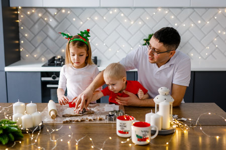 Happy family father and two kids in festive outfit making Christmas cookies together in kitchen at home during winter holidays, dad decorating xmas gingerbreads with small childrenの写真素材