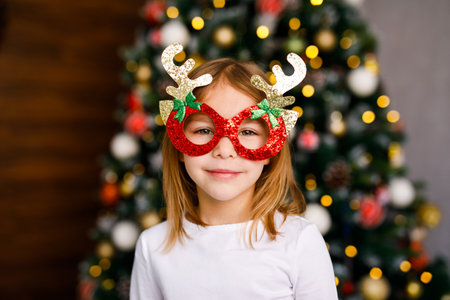 A six-year-old girl in shiny red glasses is posing against the background of a Christmas tree.の写真素材