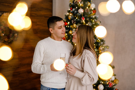A couple in love hugs and sips champagne in front of a big Christmas tree with bokeh from twinkling garland lights all around.の写真素材