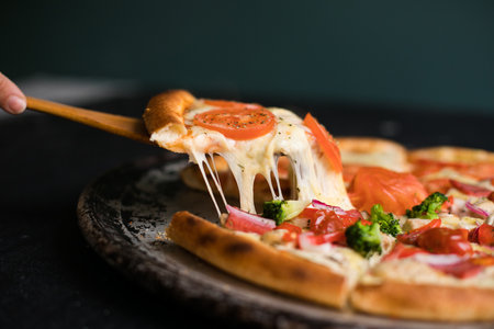 Delicious cheese stringy slice lifted of full supreme pizza baked fresh out of the oven next to ingredients.の写真素材