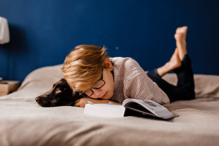 Child wearing glasses reading a book in bed with his dog.の写真素材