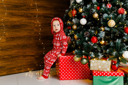 A toddler boy in a knitted pajama is sitting on a big gift under the Christmas tree.の写真素材