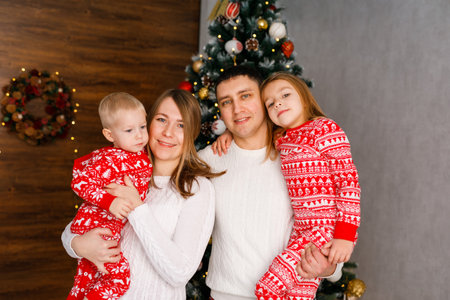 Mom, Dad, and the brother and sister in red pajamas are playing and hugging in front of the Christmas tree.の写真素材