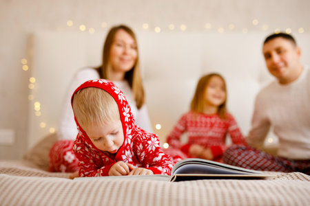 A toddler boy in a Christmas pajama is reading a book, with his parents and sister in the backgroundの写真素材