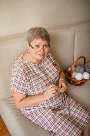 An elderly woman wearing glasses crochets at home, sitting on a beige sofa.の写真素材