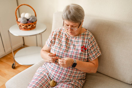 A mature woman in glasses is crocheting at home on a light beige sofa.