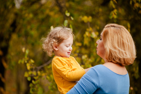 Loving mother holding toddler girl with curly hair in yellow shirt in autumn park.の写真素材