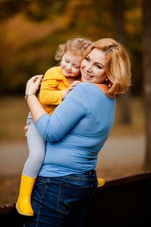 Loving mother holding toddler girl with curly hair in yellow shirt in autumn park.の写真素材