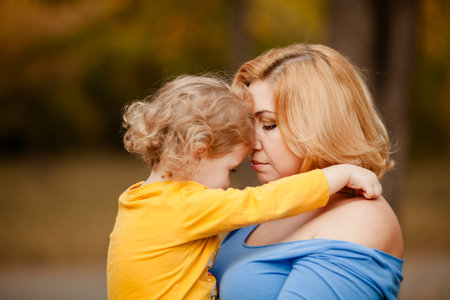 Loving mother holding toddler girl with curly hair in yellow shirt in autumn park.の写真素材