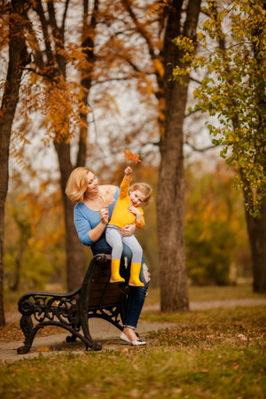 Mother holding little girl with curly hair in yellow shirt in autumn park.の写真素材