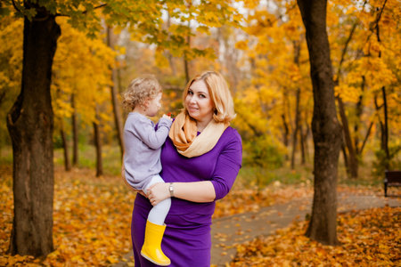 Plus-size woman in purple dress holding and playing with her 3-year-old daughter in autumn park.の写真素材