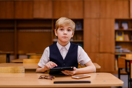 Smart schoolboy in a vest and glasses using a tablet in the school classroom.の写真素材