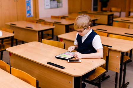 Smart schoolboy in a vest and glasses using a tablet in the school classroom.