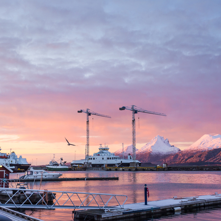 Beautiful sunrise in the port city Sandnessjoen. Berths, floating pontoons, tower cranes and moored ships on the background of the mountains and the pink sky reflected in the waters.の写真素材