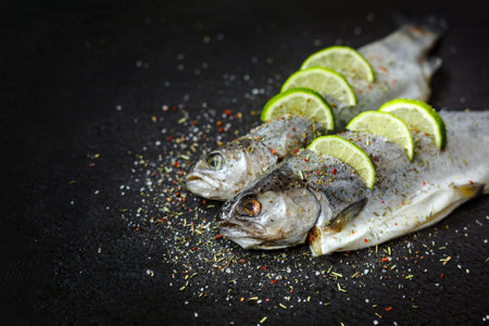 Raw fish with aromatic herbs, spices, salt and lime slices. Raw river trout on black stone tray,...