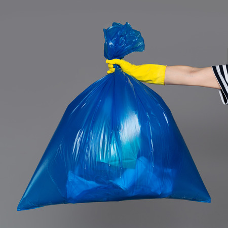 A woman's hand in a rubber glove holds a blue plastic bag full of garbage.の写真素材