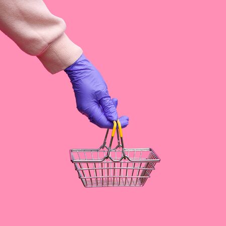 A woman's hand in a disposable bright glove holds a shopping metal basket from a supermarket on a pink background. The safety of customers during the epidemic of coronavirus.の写真素材