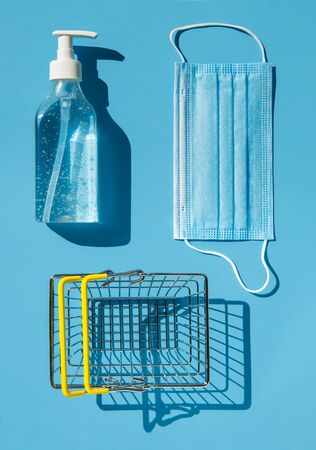 Personal protection kit for shopping during the coronavirus pandemic: face mask, gel sanitizer and shopping basket. Blue background, contrasting shadows. The view from the top.の写真素材