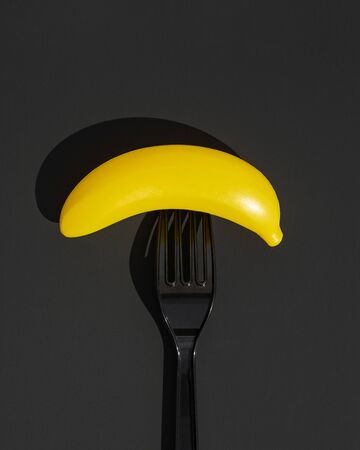 A black disposable plastic fork with a plastic banana impaled on it. The concept of ecological payback for excessive use of plastic by humans. Black background. Top view.の写真素材