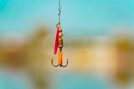 A bright spinner with a triple hook for fishing hangs on a fishing line above the water. Selective focus. Close up.の写真素材