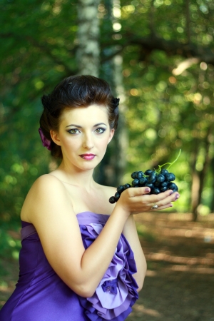 portrait of beautiful young woman in violet fashion dress holding dark blue grapesの写真素材