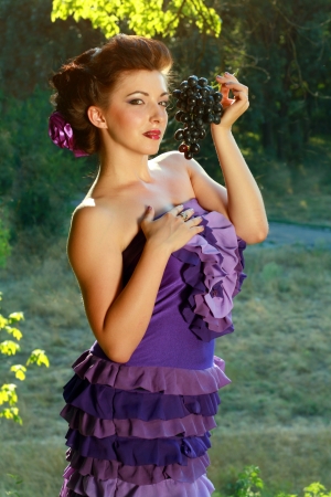 portrait of beautiful young woman in violet fashion dress holding dark blue grapesの写真素材