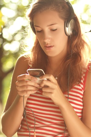 Modern happy millennial teenage girl with headphones and smartphone smiling standing outdoors in park on sunny summer day retouched, vibrant colors.の写真素材