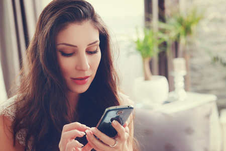 Smiling brunette sitting on her couch on a phone call at home in the living room. Retro soft toning on image warm colors.の写真素材