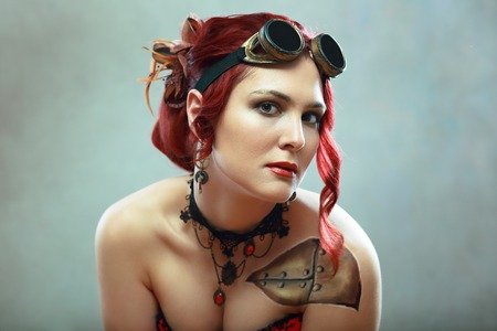 portrait of beautiful redhead steampunk woman wearing flying goggles and corset posing next to color backgroundの写真素材