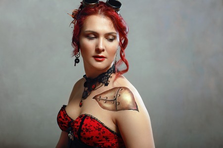 portrait of beautiful redhead steampunk woman wearing flying goggles and corset posing next to color backgroundの写真素材